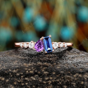 May include: A rose gold ring featuring a cluster of gemstones. The ring has a rectangular blue gemstone, a triangular purple gemstone, and several small white gemstones. The ring is set on a dark, textured surface.