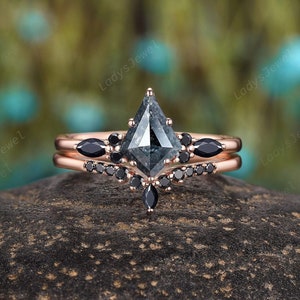 May include: A rose gold engagement ring set with a gray, kite-shaped diamond and black diamonds. The ring is set on a gray rock.