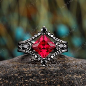 Puede incluir: Un anillo negro con una gran piedra preciosa roja vibrante de corte cuadrado en el centro. El anillo está rodeado de piedras negras más pequeñas y un intrincado trabajo de metal. El anillo está sobre una superficie oscura y texturizada.