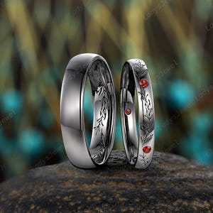 May include: Two silver wedding rings. One ring is plain, polished, and reflective. The other ring has an engraved floral pattern with three small red gemstones. The rings are displayed on a dark, textured surface.