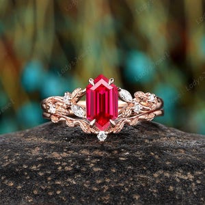 May include: A rose gold engagement ring with a large, faceted, rectangular ruby gemstone. The ring features a floral design with small, clear accent stones and leaf details. The ring is set on a dark, textured surface.