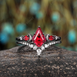 May include: A black gold ring set with a large red gemstone and smaller red gemstones on the sides. The ring has a crescent moon design on each side of the large gemstone. The ring is set on a black rock.