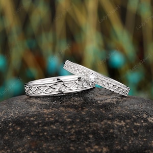 Puede incluir: Dos anillos de boda plateados con intrincados diseños de nudos celtas. Un anillo presenta un pequeño diamante redondo. Los anillos se muestran sobre una superficie oscura y texturizada, con un fondo borroso verde y dorado.