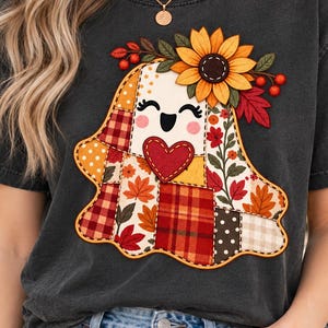 May include: A charcoal gray t-shirt featuring a central graphic of a ghost made of various autumn-themed fabric patches. The ghost has a red heart, a smiling face, and is adorned with a large yellow sunflower and red berries on its head.