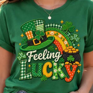 Op de afbeelding: Een groen t-shirt met een grafische print van een kabouterhoed, een regenboog, een pot met goud en klavertjes. De tekst "Feeling Lucky" is gedrukt in een gestileerd lettertype. Het ontwerp bevat ruitjes, stippen en bloemmotieven in groen, oranje en geel.