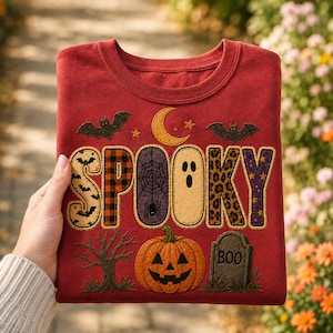 May include: A red crewneck sweatshirt featuring the word SPOOKY in large, patterned letters. The design includes a crescent moon, bats, a spider web, a ghost, a jack-o'-lantern, a bare tree, and a tombstone with the text BOO printed on it.