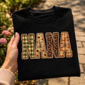 Może przedstawiać: Czarny t-shirt z krótkim rękawem z napisem NANA wykonanym dużymi literami blokowymi. Każda litera ma inny wzór: zielona kratka, słoneczniki i dynie, wzór w panterkę oraz pomarańczowa kratka w białe stokrotki, całość wykończona ozdobnym ściegiem.