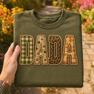 May include: An olive green crewneck t-shirt featuring the word DADA in large block letters. Each letter has a unique pattern: the first is green gingham, the second has pumpkins and sunflowers, the third is leopard print, and the fourth is orange plaid with white daisies.