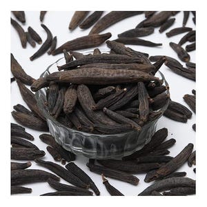 May include: A glass bowl containing numerous dark brown, elongated dried pods, scattered on a white surface. The pods have a textured appearance, possibly a spice or herb, and are a deep brown colour.