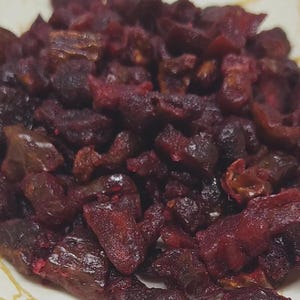 May include: A close-up shot of a pile of dried, dark red fruit pieces. The pieces have a wrinkled texture and varying shapes, with some appearing more elongated and others rounder. The fruit is likely a type of dried berry or cherry.