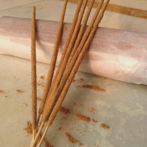 Puede incluir: Primer plano de varios palitos de incienso con un revestimiento marr&oacute;n texturizado. Los palitos est&aacute;n dispuestos en forma de abanico, con algunos gr&aacute;nulos dispersos. En el fondo se ve un paquete de papel enrollado.