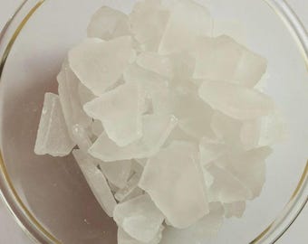 Puede incluir: Un cuenco de vidrio transparente lleno de cristales blancos translúcidos de forma irregular. Los cristales tienen un aspecto ligeramente esmerilado y varían en tamaño, creando una superficie texturizada y desigual. El borde del cuenco es visible.