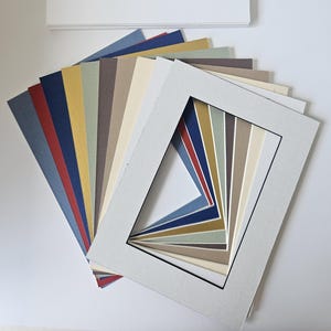 May include: A selection of coloured mount boards and a stack of white paper. The mount boards are fanned out, displaying a variety of colours such as blue, red, yellow, and neutral shades. The white paper is neatly stacked.