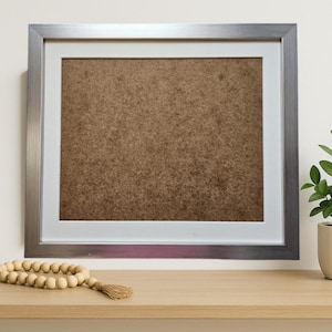 May include: A silver framed picture frame with a white mount and brown backing sits on a light wood shelf. A small potted plant with green leaves and a wooden bead garland with a tassel are also on the shelf.