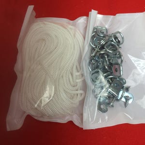 May include: Two clear plastic bags containing hardware. One bag holds a coiled white rope. The other bag contains metal brackets, screws, and rings. The items are on a red surface.