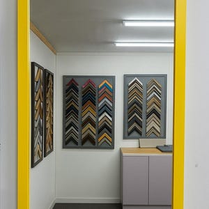 May include: A large rectangular mirror with a bright yellow frame. The mirror reflects a room with framed samples of picture frames in various colours and styles, displayed on the walls. The room has a grey carpet and a grey and wood-toned cabinet.