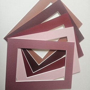 May include: A selection of rectangular picture mounts in shades of pink and burgundy. The mounts are arranged in a fan shape, revealing a layered pattern of colours through the openings. These mounts are designed for framing artwork or photographs.