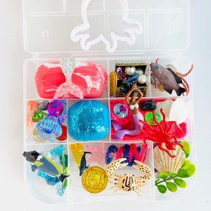 Puede incluir: Una caja de almacenamiento de pl&aacute;stico transparente con tem&aacute;tica de sirena. La caja contiene una variedad de juguetes, incluyendo una mu&ntilde;eca sirena, un cofre del tesoro, un cangrejo, una estrella de mar, una concha y una esfera azul. La caja est&aacute; dividida en compartimentos y tiene un recorte de pulpo blanco en la tapa.