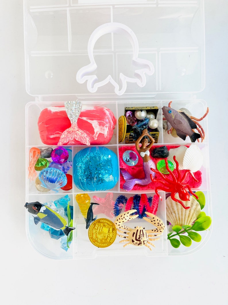 Puede incluir: Una caja de almacenamiento de pl&aacute;stico transparente con tem&aacute;tica de sirena. La caja contiene una variedad de juguetes, incluyendo una mu&ntilde;eca sirena, un cofre del tesoro, un cangrejo, una estrella de mar, una concha y una esfera azul. La caja est&aacute; dividida en compartimentos y tiene un recorte de pulpo blanco en la tapa.