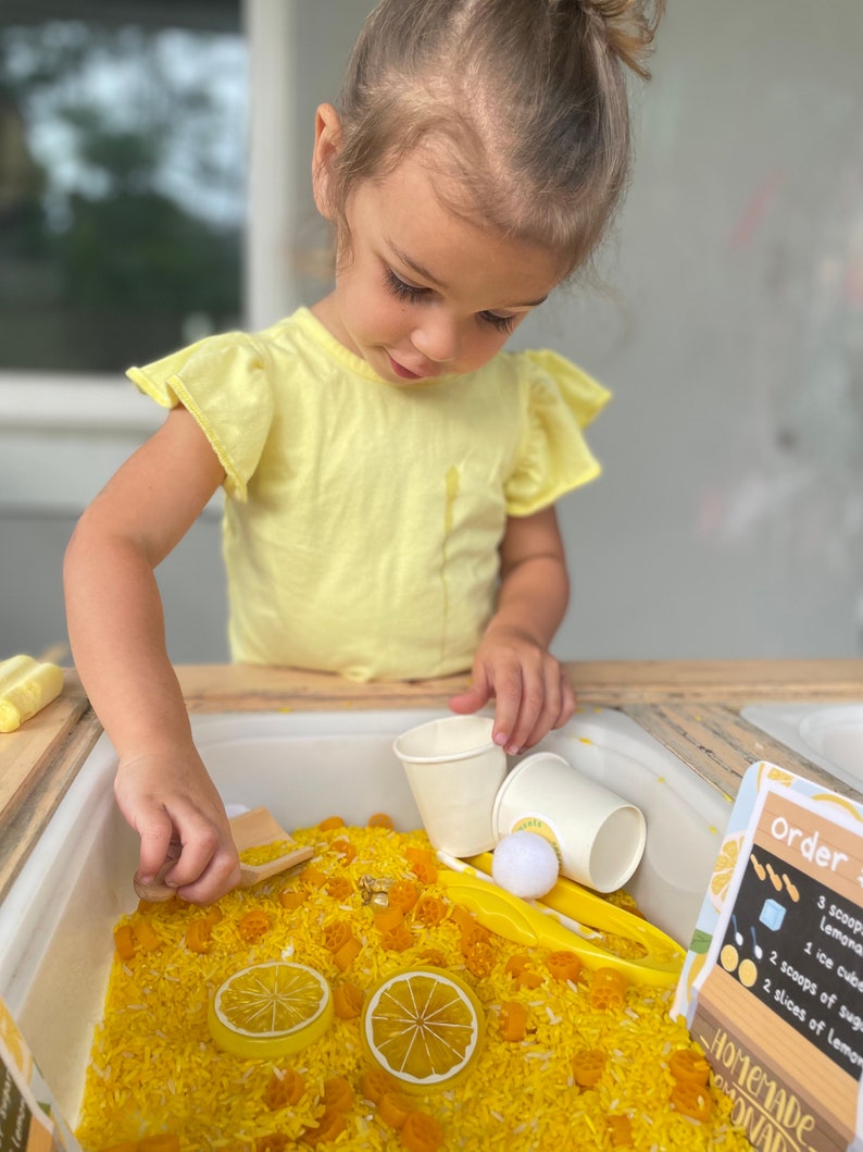 Puede incluir: Una ni&ntilde;a con una camisa amarilla juega con un recipiente sensorial lleno de arroz amarillo, rodajas de lim&oacute;n y otros juguetes. El recipiente incluye tazas, cucharas y un letrero con la frase "Homemade Lemonade".