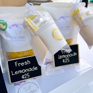 Puede incluir: Exhibici&oacute;n de kits de juego sensorial "Fresh Lemonade". Cada kit incluye una taza blanca, un mortero de madera, pajitas amarillas y blancas y cubitos de hielo transparentes. Los kits est&aacute;n empaquetados en bolsas transparentes con el logo "Little Elements".