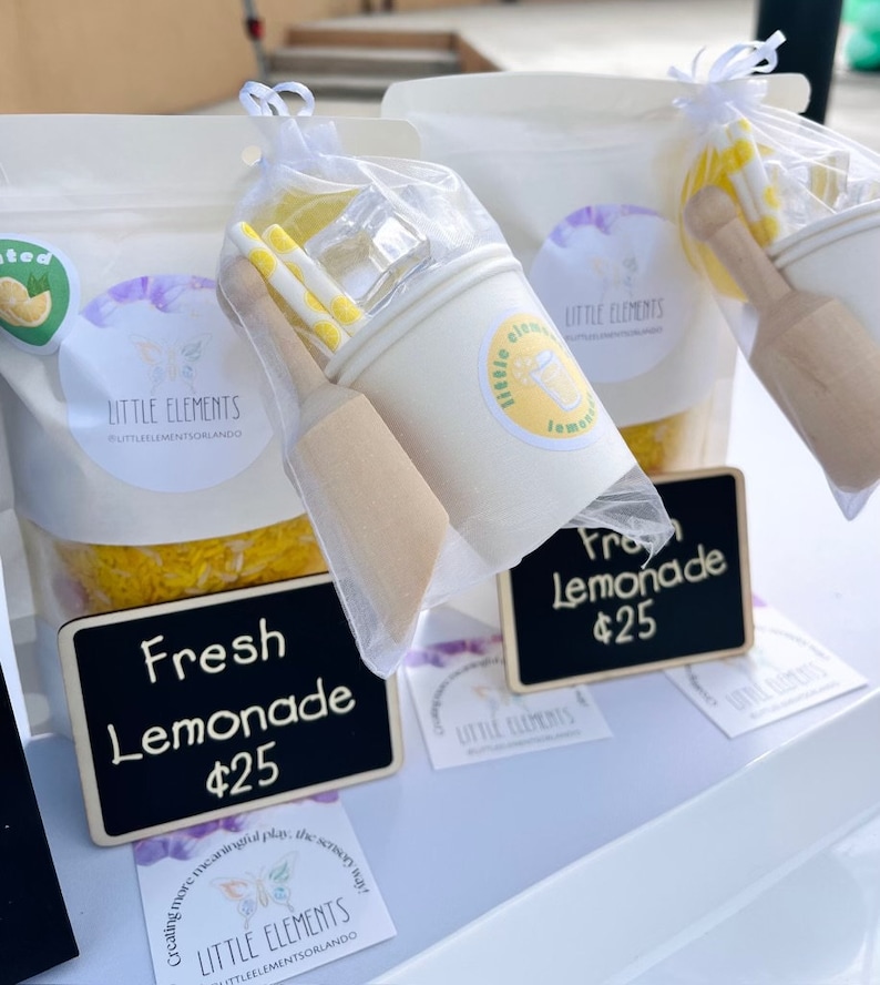 Puede incluir: Exhibici&oacute;n de kits de juego sensorial "Fresh Lemonade". Cada kit incluye una taza blanca, un mortero de madera, pajitas amarillas y blancas y cubitos de hielo transparentes. Los kits est&aacute;n empaquetados en bolsas transparentes con el logo "Little Elements".