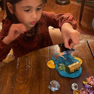 Puede incluir: Una ni&ntilde;a juega con una figura de sirena, un cofre del tesoro y monedas de oro sobre una superficie de plastilina azul. La plastilina est&aacute; rodeada de gemas de colores y una concha.