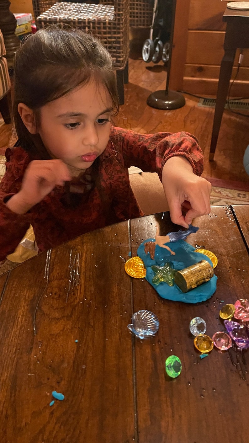 Puede incluir: Una ni&ntilde;a juega con una figura de sirena, un cofre del tesoro y monedas de oro sobre una superficie de plastilina azul. La plastilina est&aacute; rodeada de gemas de colores y una concha.