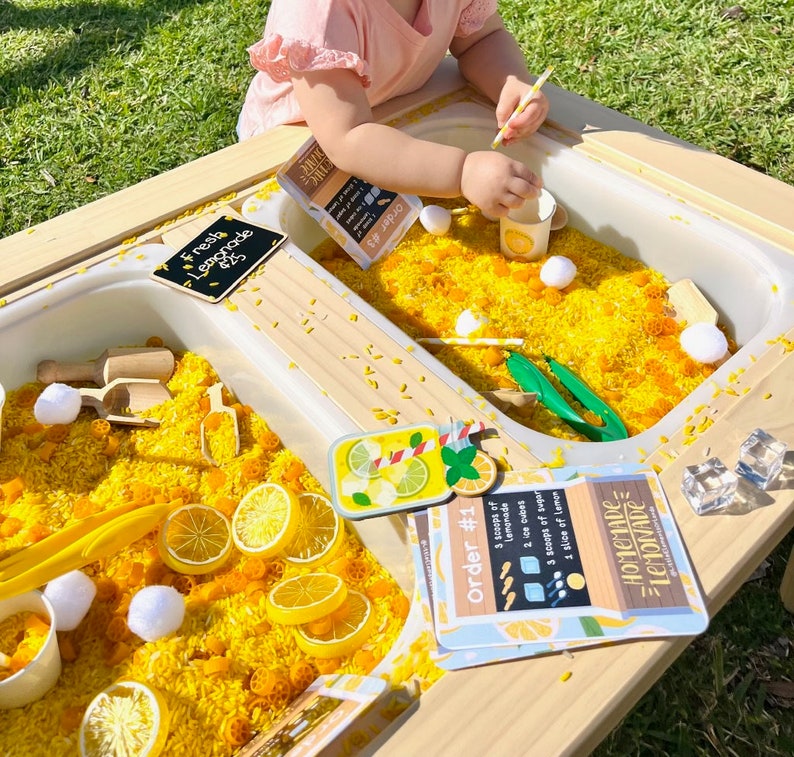 Puede incluir: Una mesa sensorial de madera llena de arroz amarillo, rodajas de lim&oacute;n y accesorios de juego. La mesa incluye letreros que dicen "Fresh Lemonade" y "Homemade Lemonade". Tambi&eacute;n hay tazas peque&ntilde;as, pinzas y otros art&iacute;culos de juego.