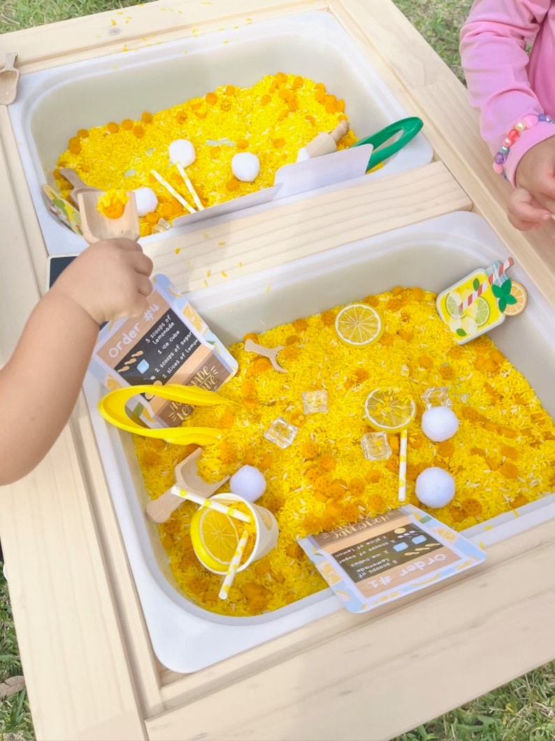 Puede incluir: Un juego sensorial con arroz te&ntilde;ido de amarillo, cucharas de madera y peque&ntilde;as bolas blancas. El juego incluye tarjetas impresas con recetas de limonada y cubitos de hielo de pl&aacute;stico. La mano de un ni&ntilde;o sostiene una cuchara.