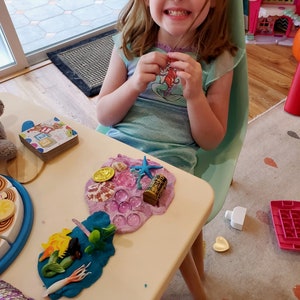 Puede incluir: Una ni&ntilde;a juega con un juego de plastilina con tem&aacute;tica de sirena. El juego incluye una figura de sirena, una estrella de mar, una concha y un cofre del tesoro. La plastilina es rosa y morada y est&aacute; extendida sobre una mesa blanca.