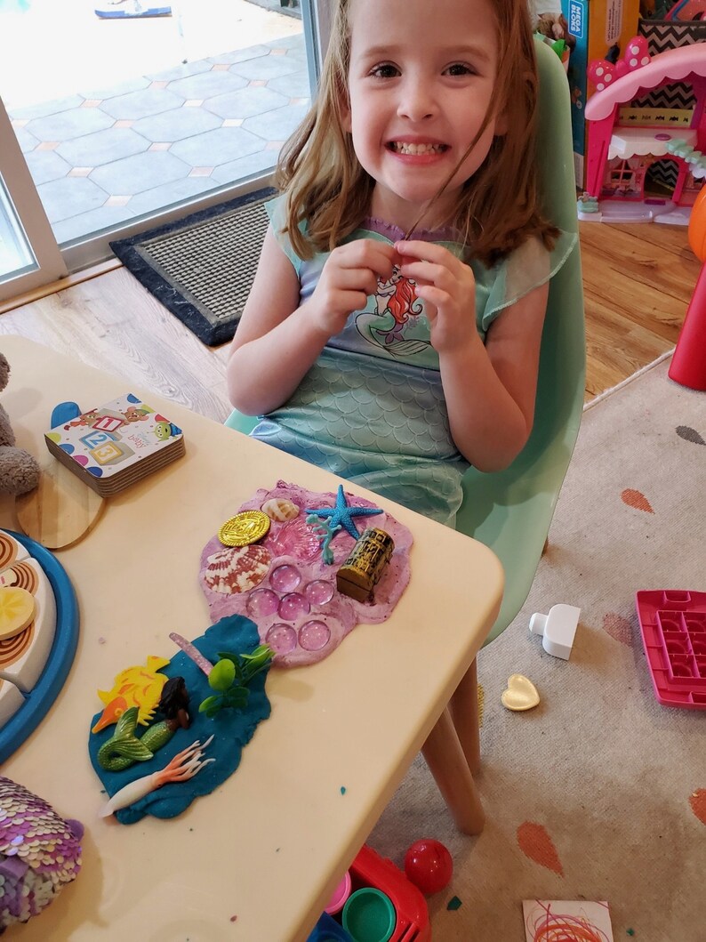 Puede incluir: Una ni&ntilde;a juega con un juego de plastilina con tem&aacute;tica de sirena. El juego incluye una figura de sirena, una estrella de mar, una concha y un cofre del tesoro. La plastilina es rosa y morada y est&aacute; extendida sobre una mesa blanca.