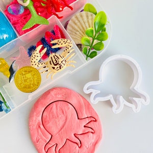 Puede incluir: Un cortador de galletas de pl&aacute;stico blanco en forma de pulpo con una impresi&oacute;n de plastilina rosa en forma de pulpo a su lado. El cortador de galletas est&aacute; sobre una superficie blanca.