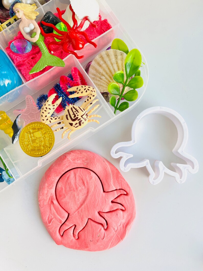Puede incluir: Un cortador de galletas de pl&aacute;stico blanco en forma de pulpo con una impresi&oacute;n de plastilina rosa en forma de pulpo a su lado. El cortador de galletas est&aacute; sobre una superficie blanca.