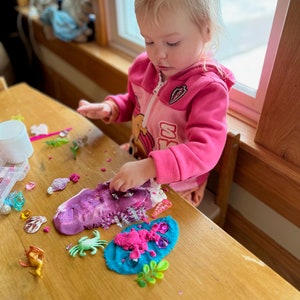 Puede incluir: Una ni&ntilde;a peque&ntilde;a juega con plastilina rosa, azul y morada en una mesa de madera. La plastilina est&aacute; decorada con purpurina, criaturas marinas de pl&aacute;stico y otros juguetes peque&ntilde;os.