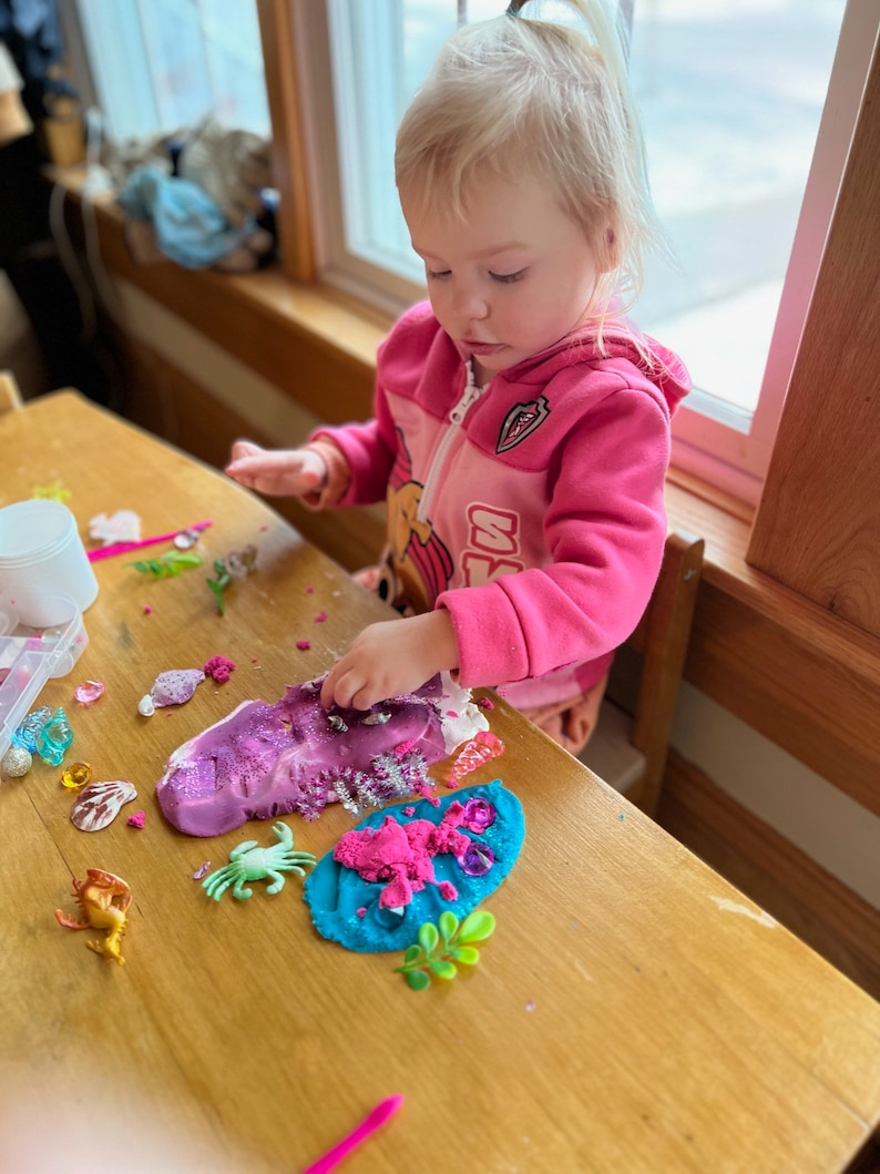 Puede incluir: Una ni&ntilde;a peque&ntilde;a juega con plastilina rosa, azul y morada en una mesa de madera. La plastilina est&aacute; decorada con purpurina, criaturas marinas de pl&aacute;stico y otros juguetes peque&ntilde;os.