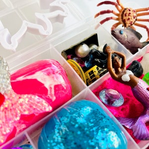 Puede incluir: Una caja de almacenamiento de pl&aacute;stico transparente llena de varios juguetes y accesorios con tem&aacute;tica de sirenas. La caja incluye slime rosa y azul, una figura de sirena, un cangrejo, un cofre del tesoro y gemas.