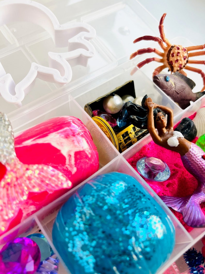 Puede incluir: Una caja de almacenamiento de pl&aacute;stico transparente llena de varios juguetes y accesorios con tem&aacute;tica de sirenas. La caja incluye slime rosa y azul, una figura de sirena, un cangrejo, un cofre del tesoro y gemas.