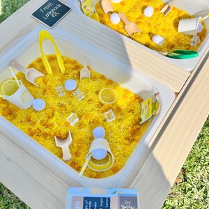 Puede incluir: Una mesa sensorial de madera con dos cubos blancos llenos de arroz amarillo, cucharas de madera y accesorios con tem&aacute;tica de lim&oacute;n. La mesa incluye un letrero que dice "Fresh Lemonade" y otro con una receta de limonada.