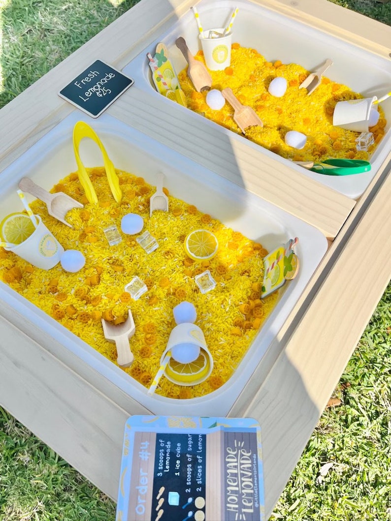 Puede incluir: Una mesa sensorial de madera con dos cubos blancos llenos de arroz amarillo, cucharas de madera y accesorios con tem&aacute;tica de lim&oacute;n. La mesa incluye un letrero que dice "Fresh Lemonade" y otro con una receta de limonada.