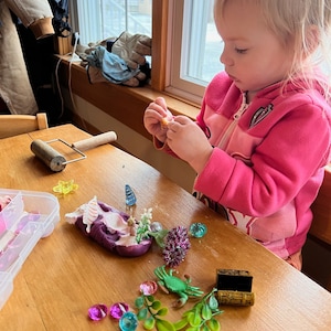 Puede incluir: Un ni&ntilde;o juega con una creaci&oacute;n de plastilina morada, rodeada de gemas de colores, un cangrejo verde y un peque&ntilde;o cofre del tesoro de madera. La creaci&oacute;n de plastilina presenta una concha blanca y una peque&ntilde;a flor.