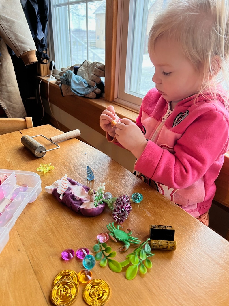 Puede incluir: Un ni&ntilde;o juega con una creaci&oacute;n de plastilina morada, rodeada de gemas de colores, un cangrejo verde y un peque&ntilde;o cofre del tesoro de madera. La creaci&oacute;n de plastilina presenta una concha blanca y una peque&ntilde;a flor.
