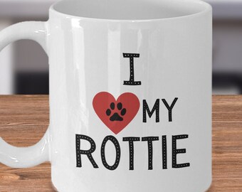 May include: White ceramic mug with the text "I ❤️ MY ROTTIE". The heart is red with a black paw print inside. The mug has a curved handle and is on a wooden surface.