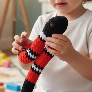 May include: A handmade crochet snake toy in black, red, and white. The toy has a black head with red eyes and a red and white striped body. The snake is being held by a child.