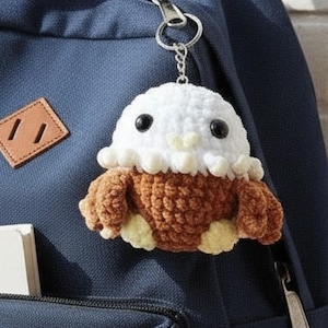 May include: A handmade crochet keychain featuring a white and brown creature with black eyes. The keychain is attached to a silver keyring and hangs from a navy blue backpack. The creature has a fluffy white head and brown body.