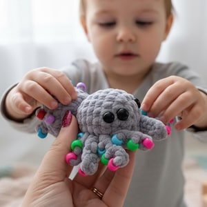May include: A gray crocheted octopus toy with large black eyes and colorful beads on its tentacles. The octopus is being held by a person and a child. The tentacles have pink, green, blue, and purple beads.