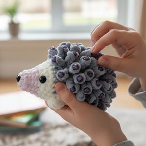May include: A handmade crochet hedgehog toy. The toy features a gray body with purple bead accents, a white and light pink face, and black eyes. The soft, plush toy is held in someone's hands.