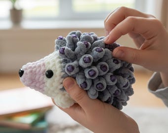 May include: A handmade crochet hedgehog toy. The toy features a gray body with purple bead accents, a white and light pink face, and black eyes. The soft, plush toy is held in someone's hands.