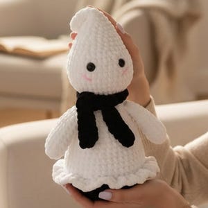 Op de afbeelding: Een wit gehaakt spook knuffeltje met een zachte, getextureerde garen afwerking, twee kleine zwarte kraaloogjes en subtiel roze geborduurde wangen. Het spookje draagt een dikke, zwarte gehaakte sjaal om de nek en heeft een geplooide onderkant.