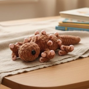 May include: A brown crocheted lizard plush toy with pink accents on its feet and back. The toy has a black eye and is lying on a beige cloth. The lizard is a handmade item.
